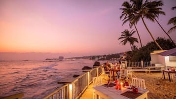 Un restaurant situé en bord de mer, avec une terrasse offrant une vue sur l’océan, les palmiers et le coucher du soleil.