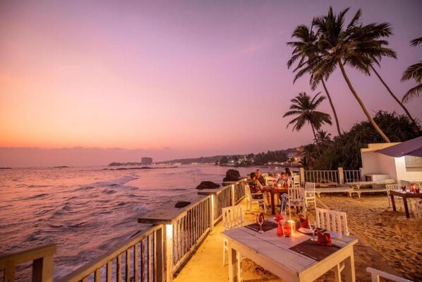 A beachside restaurant with outdoor seating overlooking the ocean, palm trees, and a sunset view.