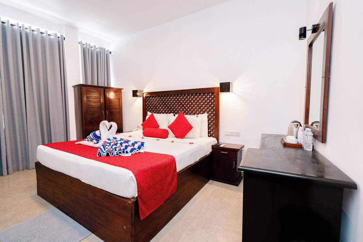 A hotel room with a wooden bed, red and white bedding, a small bedside table, a nightstand, a mirror, and a desk with a chair.