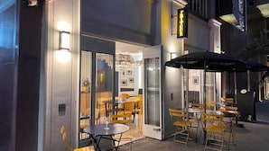 Un café al aire libre con mesas y sillas, un acceso por puerta de vidrio y un letrero sobre la entrada.
