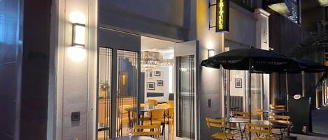 An outdoor café with tables and chairs, a glass door entrance, and a sign above the door.