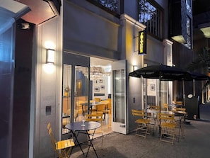 Un café en plein air avec des tables et des chaises, une entrée avec une porte vitrée et une enseigne au-dessus de la porte.