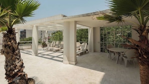 Un área de estar al aire libre con sillas y mesas, rodeada de palmeras y una estructura cubierta.