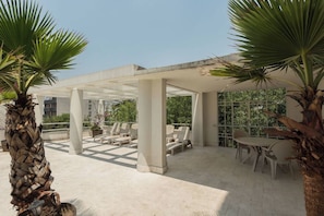 Un espace extérieur aménagé avec des chaises et des tables, entouré de palmiers et abrité par une structure couverte.
