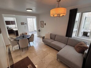 A modern living room with a sofa, dining table, and chairs. There is a chandelier, a fan, and a window with curtains.