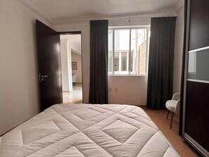 A bedroom with a large bed, a window with curtains, a door leading to another room, and a chair.