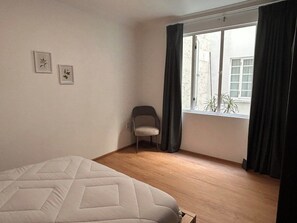 Une pièce comprenant un lit, une chaise, une fenêtre avec des rideaux et deux tableaux encadrés accrochés au mur.