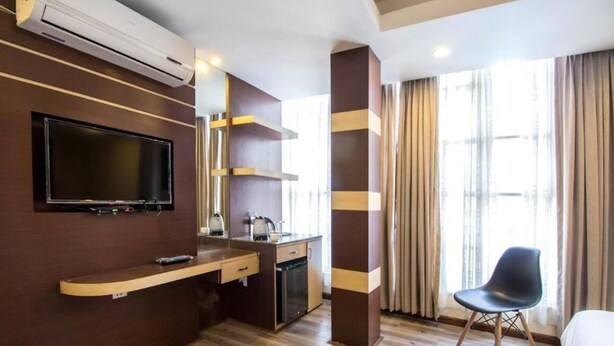 A hotel room with a flat-screen TV mounted on the wall, a wooden desk with a chair, a small kitchenette, and a large window with curtains. - Cosy Stay & Dine (Kathmandu)