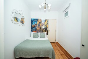 Quarto com cama, espelho, pintura de Lisboa e uma porta.