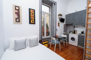 A compact living space with a bed, a small table, a chair, a washing machine, and a kitchen area. - Studio Apartment Located Central Lisbon (Lisbon)