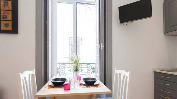 A dining area with a wooden table, two white chairs, a potted plant, and a TV mounted on the wall. - Studio Apartment Located Central Lisbon (Lisbon)