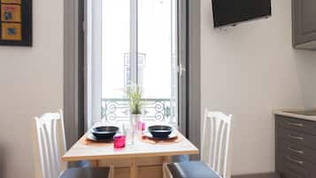 Uma área de refeições com uma mesa de madeira, duas cadeiras brancas, uma planta em vaso e uma TV fixada na parede.