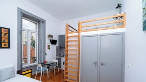 A compact room with a bed, a desk, a washing machine, a wardrobe, and a window with a view of a building. - Studio Apartment Located Central Lisbon (Lisbon)