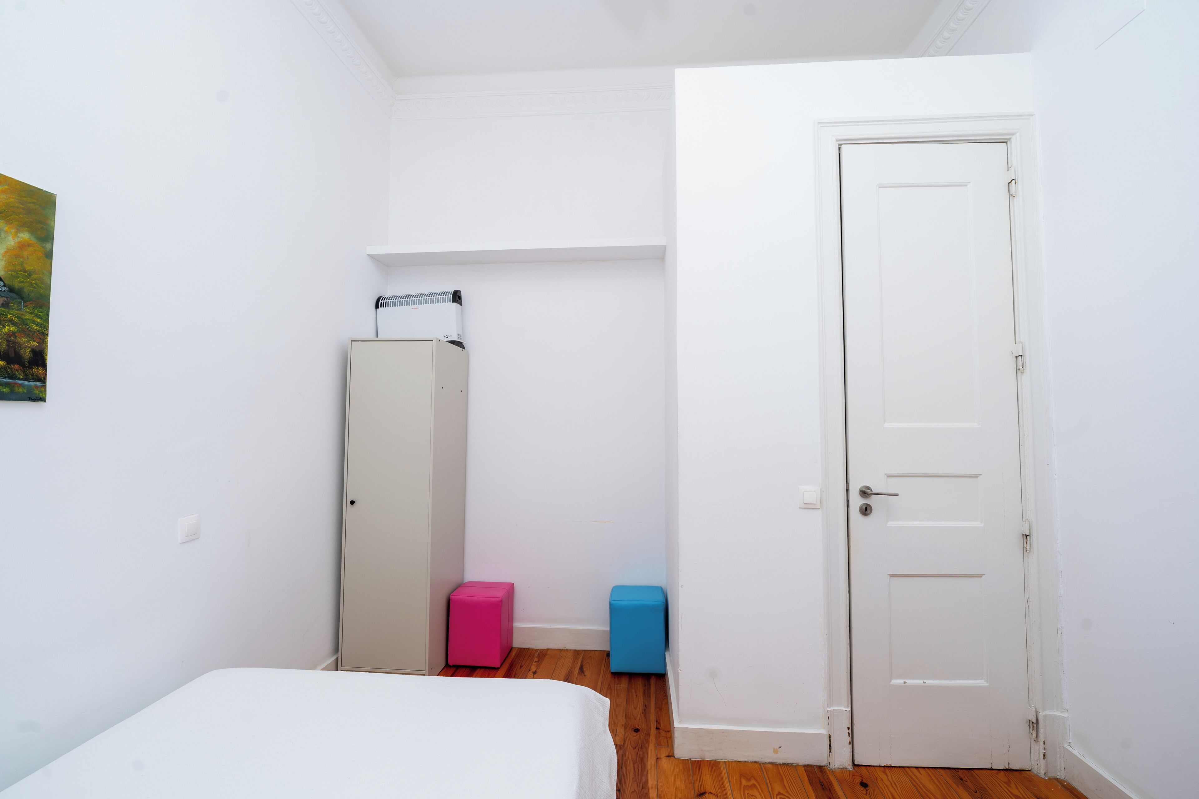 Ein kleiner Raum mit einem Bett, einer weißen Tür, einer weißen Wand und einem Holzboden.
