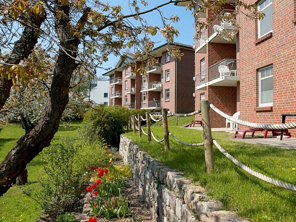 Un edificio de ladrillo con varios balcones, un muro de piedra de contención y un área verde con flores.