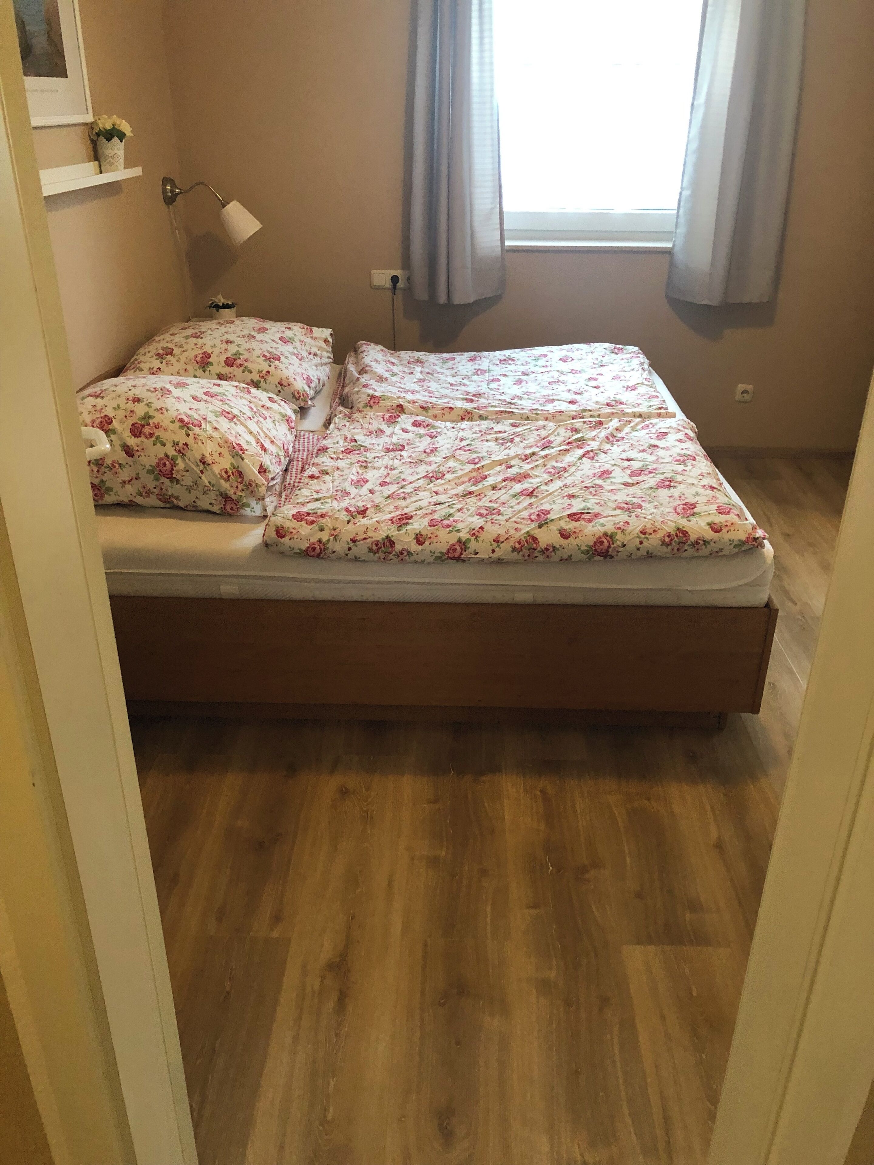 A bedroom with a wooden floor, a bed with floral bedding, a bedside lamp, and a window with curtains.