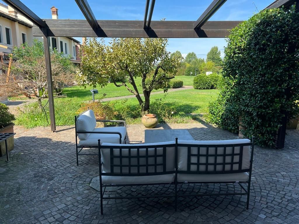 Un espace extérieur abrité avec deux chaises et une table, un jardin avec un arbre, et une vue sur un bâtiment en arrière-plan.