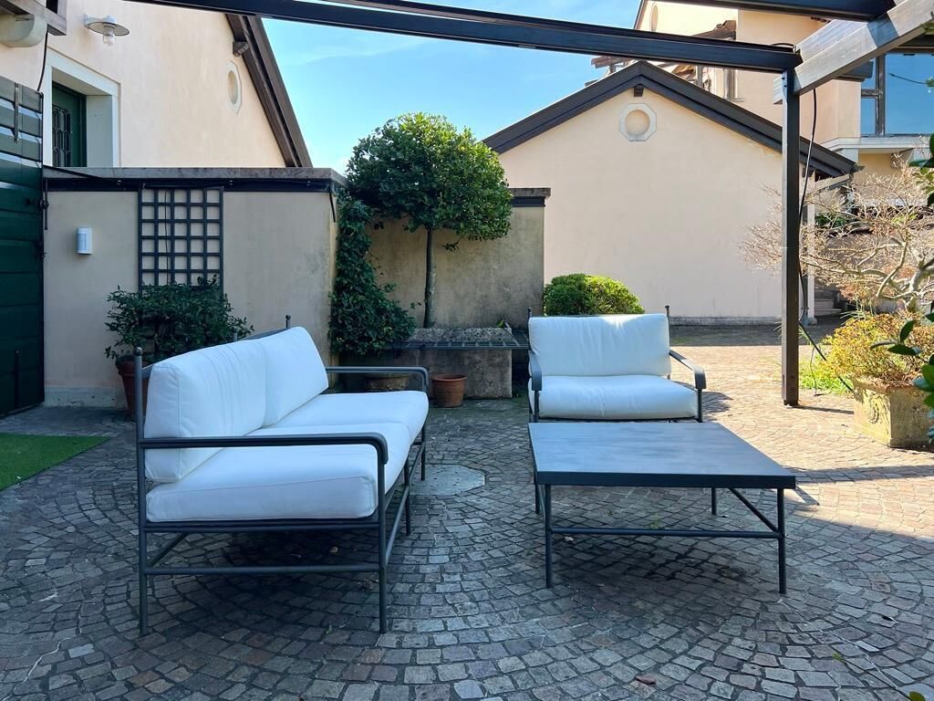 A courtyard with two white cushioned sofas and a metal table, surrounded by cobblestone flooring and potted plants.