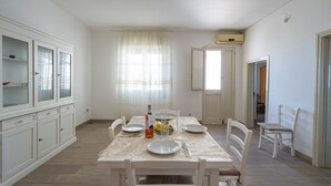 A dining area with a table set for four, white chairs, a cabinet with glass doors, and a window with sheer curtains. - Onde Dorate - Apartment in the Centre, Close to Beach (Porto Cesareo)