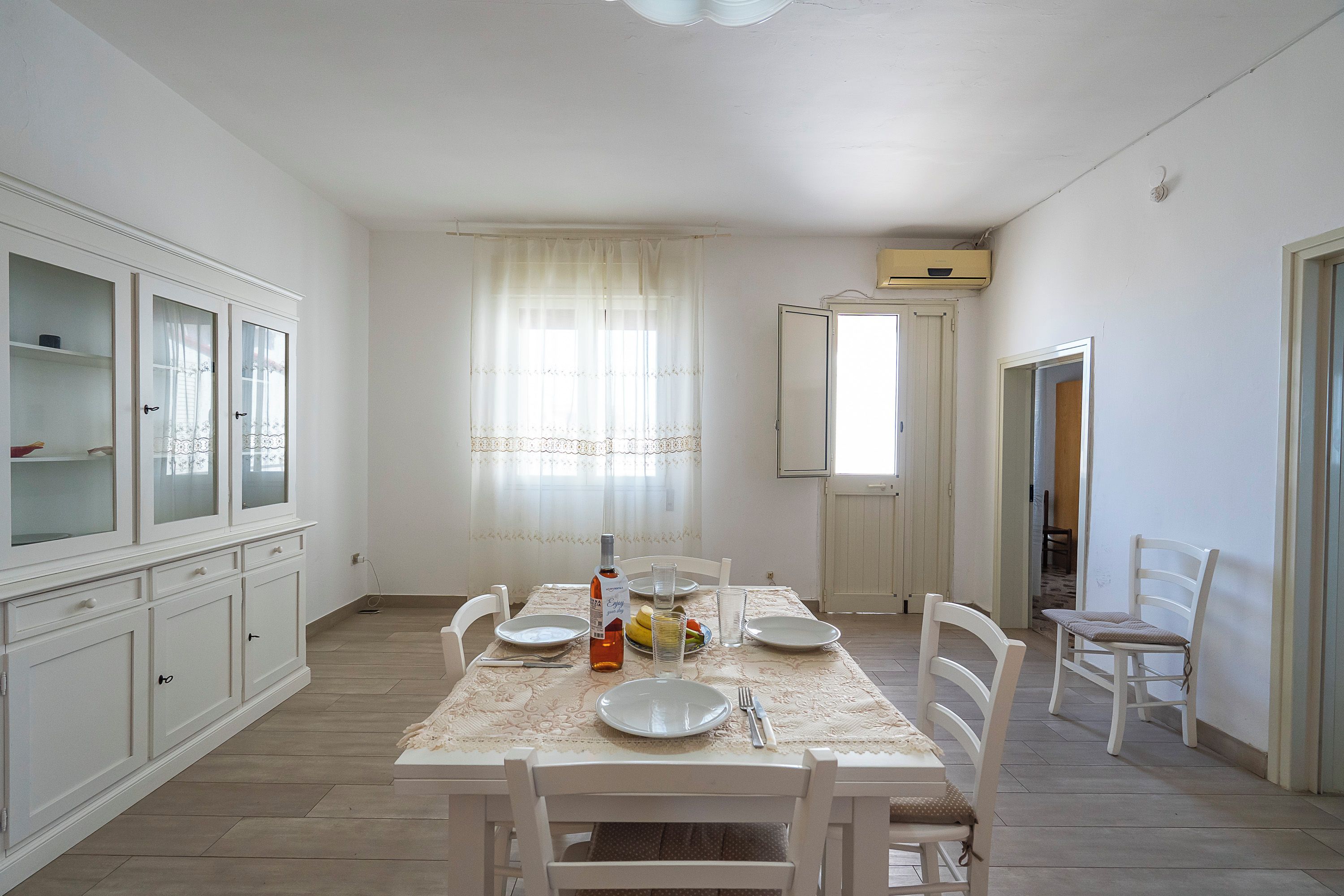 A dining area with a table set for four, white chairs, a cabinet with glass doors, and a window with sheer curtains.