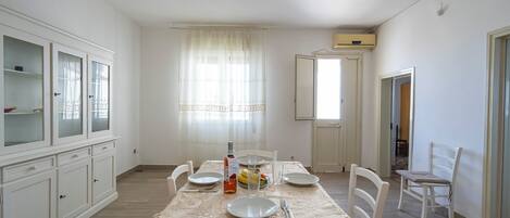 A dining area with a table set for four, white chairs, a cabinet with glass doors, and a window with sheer curtains.