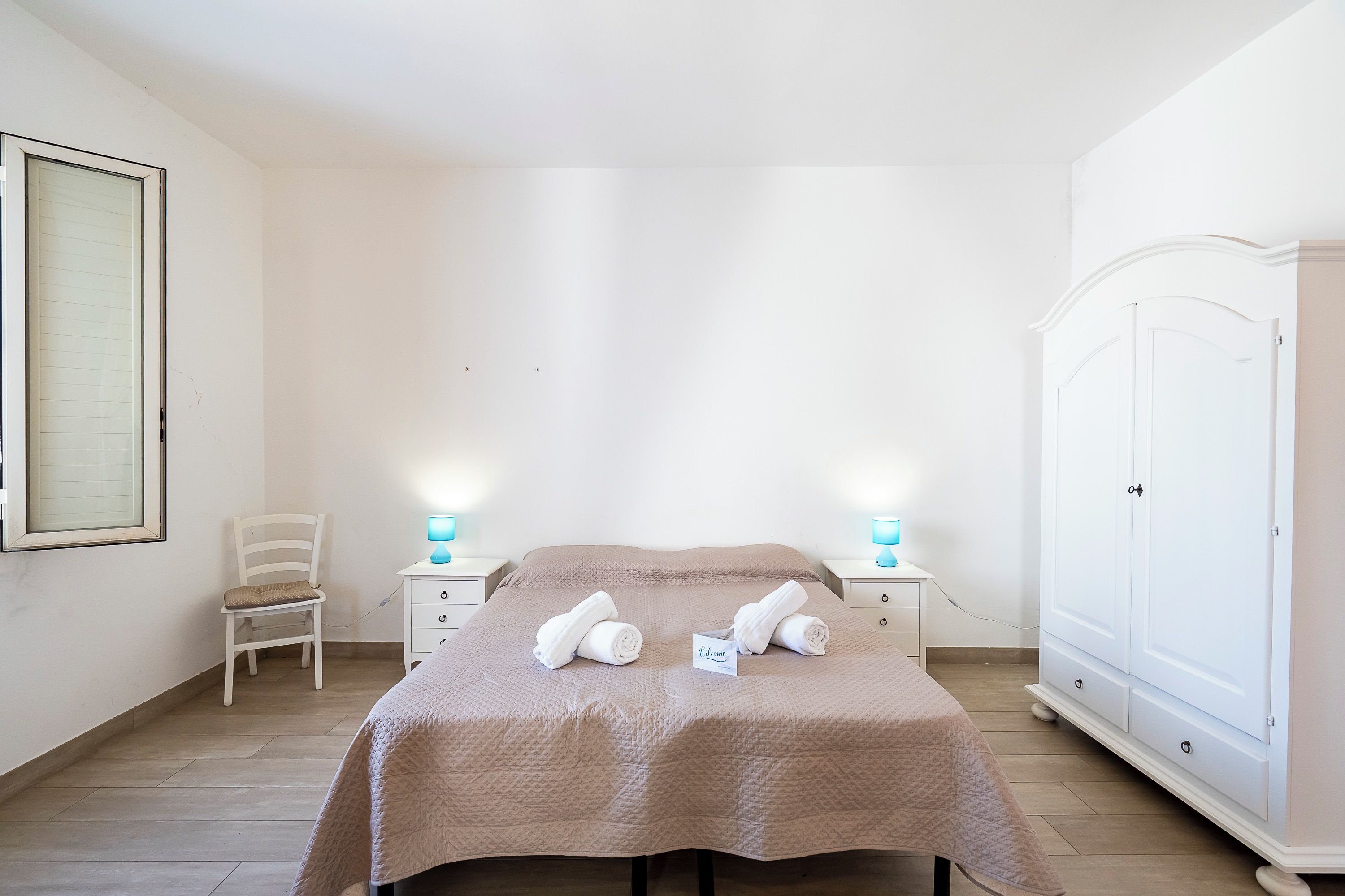 Quarto de hotel com cama, duas mesas de cabeceira, um guarda-roupa, uma cadeira e um quadro na parede.