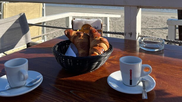 Et bord med en kurv croissanter, to hvide kopper med underkopper og en glasaskebæger på et trædæk med udsigt over en strand og hav.
