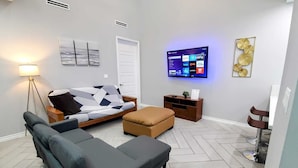 A modern living room with a sofa, ottoman, and a flat-screen TV mounted on the wall displaying various streaming services.