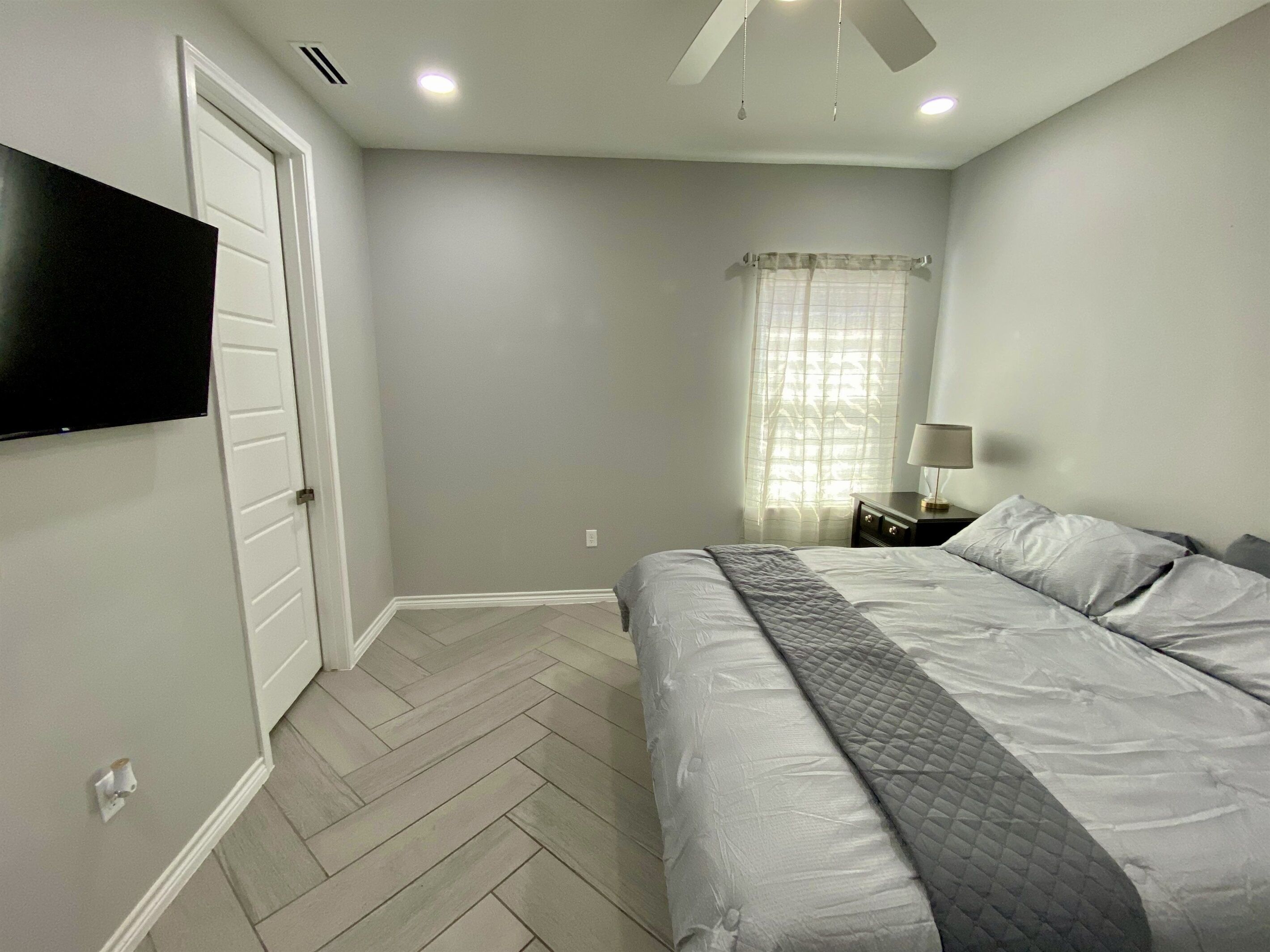 A bedroom with a bed, a TV mounted on the wall, a ceiling fan, and a window with curtains.
