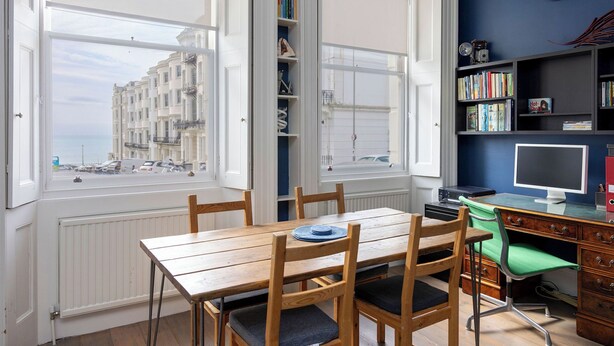 Ein Zimmer mit Esstisch, Stühlen, einem Bücherregal, einem Schreibtisch mit Computer und Blick auf Gebäude durch große Fenster.