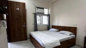 A bedroom with a wooden bed, a ceiling fan, a window with curtains, and a door with a number plate. - Sunita Residency (Gurugram)