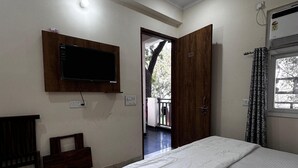 A bedroom with a bed, a TV mounted on the wall, a wooden bench, and a door leading to an outdoor area. - Sunita Residency (Gurugram)