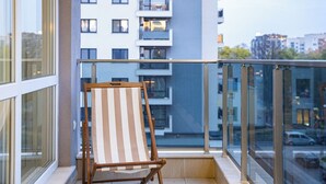 Een balkon met een houten stoel, een glazen balustrade en uitzicht op een modern gebouw met meerdere balkons.