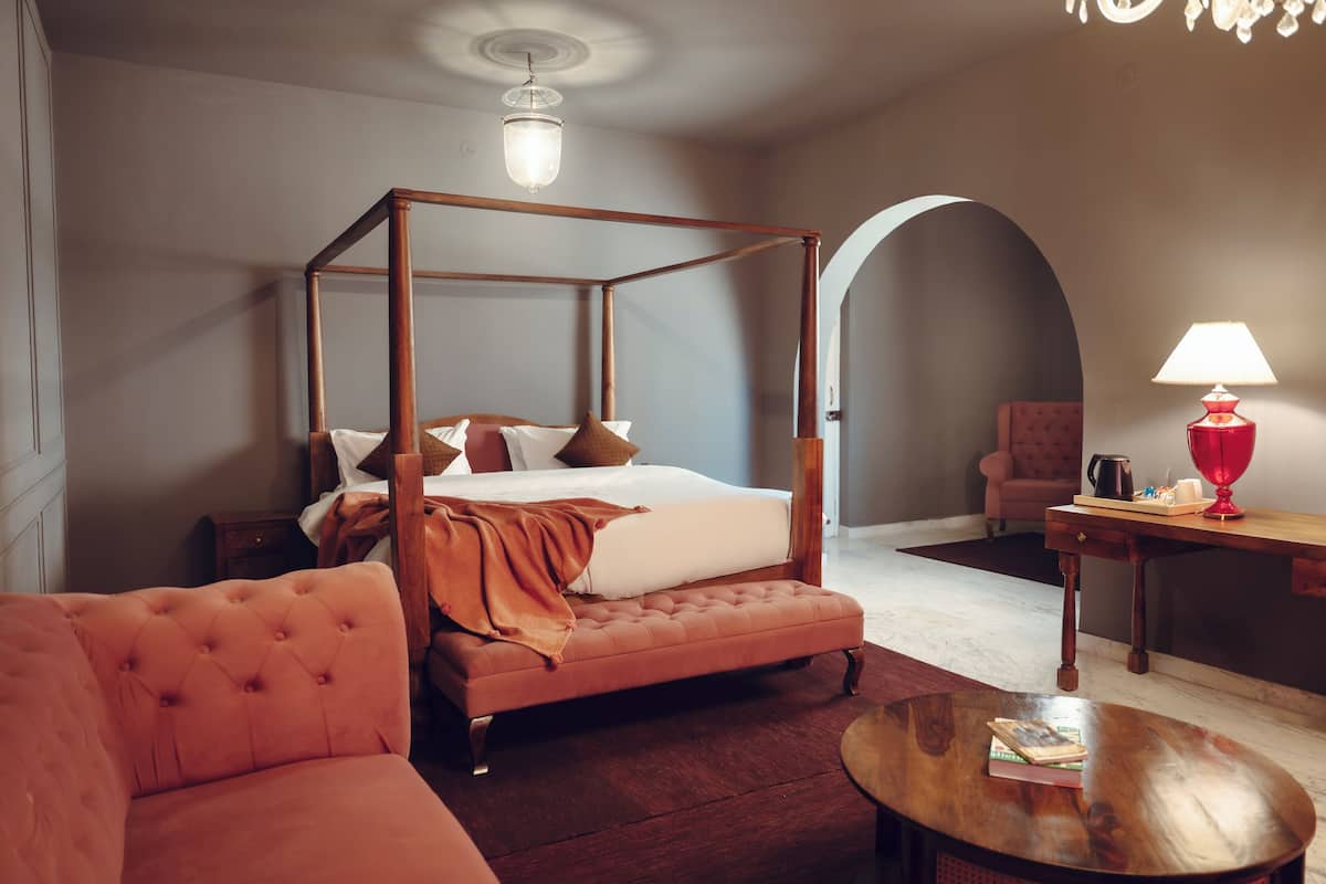 Una cama con dosel de madera, un sofá capitonado, una mesa de centro redonda de madera y una lámpara roja sobre una mesita de noche de madera.