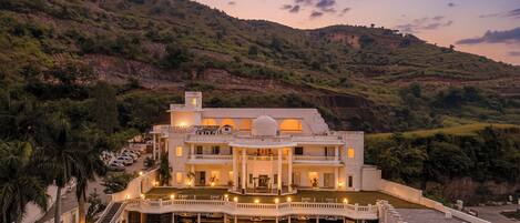 A large, multi-story building with a central courtyard, surrounded by lush greenery and hills.
