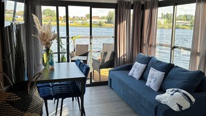 Una sala moderna con un ventanal grande que da a una masa de agua, un comedor con mesa y sillas, y un sofá azul con cojines decorativos.