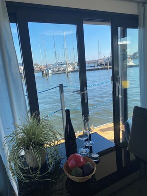 Ein Zimmer mit großem Fenster und Blick auf eine Marina mit Booten. Ein Tisch mit einer Weinflasche, Gläsern und einer Obstschale.