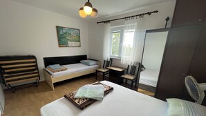 Una habitación pequeña y funcional con cama, escritorio, silla, mesita, radiador, ventana con cortinas y un cuadro en la pared.