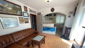 Una sala de estar con un sofá de cuero marrón, una mesa de centro de madera y un cuadro que representa una playa. Se ve un área de cocina al fondo, con armarios y una mesa de comedor.