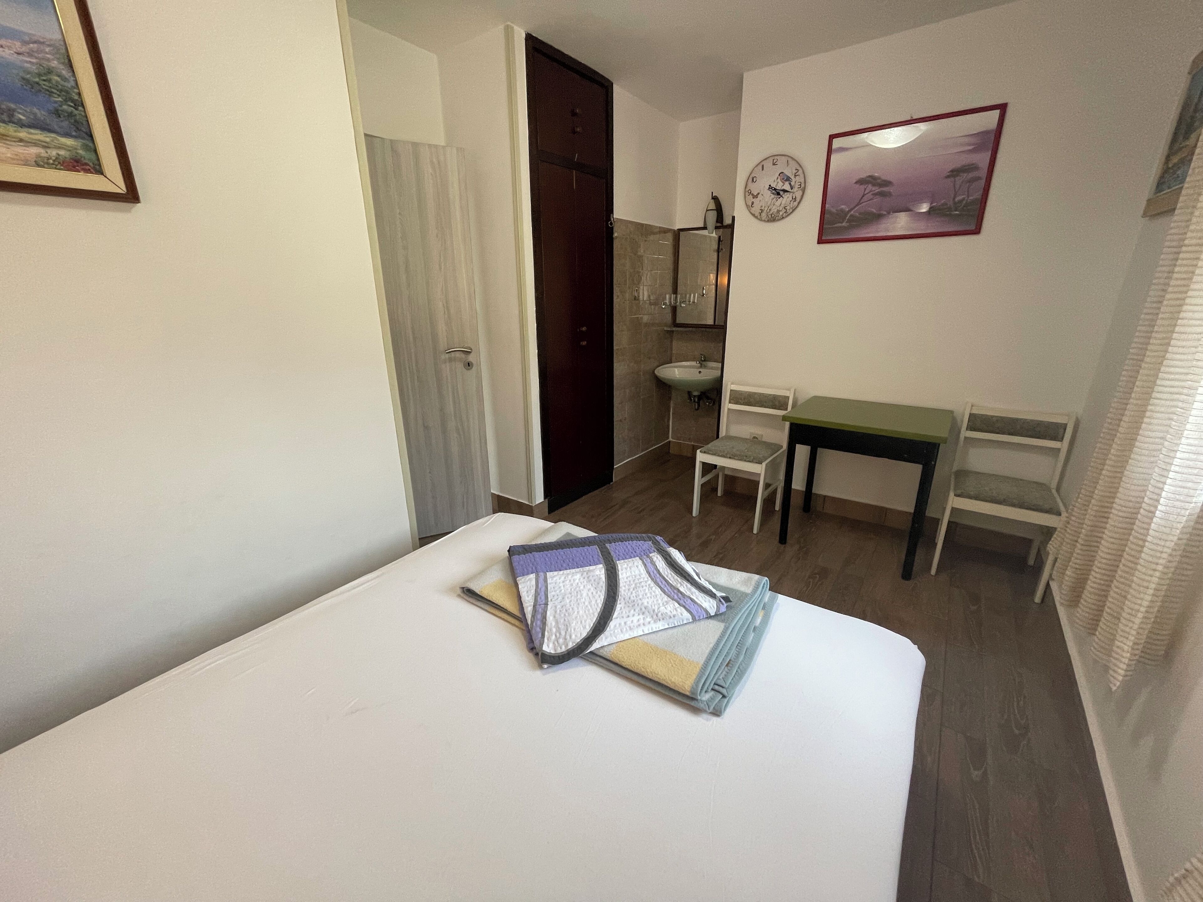 Ein Hotelzimmer mit einem Bett, einem Schreibtisch, einem Sessel, einem kleinen Tisch, einem Waschbecken und einer Uhr an der Wand.