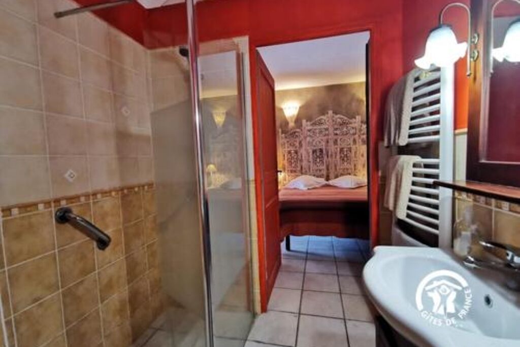 Een badkamer met een douchecabine van glas, een wastafel en een bed dat door een open deur zichtbaar is.