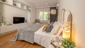 Een moderne slaapkamer met een groot bed, een televisie op een witte console, een raam met gordijnen en een bedlampje.