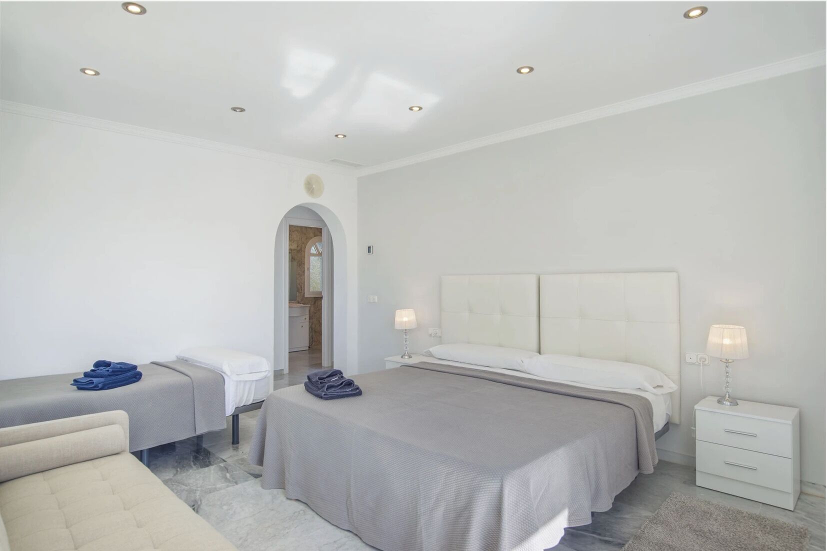 A modern hotel room with a large bed, a bedside table with a lamp, a small seating area with a chair, and a bathroom visible through an archway.