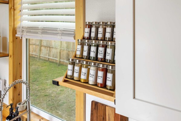 A kitchen with a window, a wooden shelf with jars of spices, and a faucet.