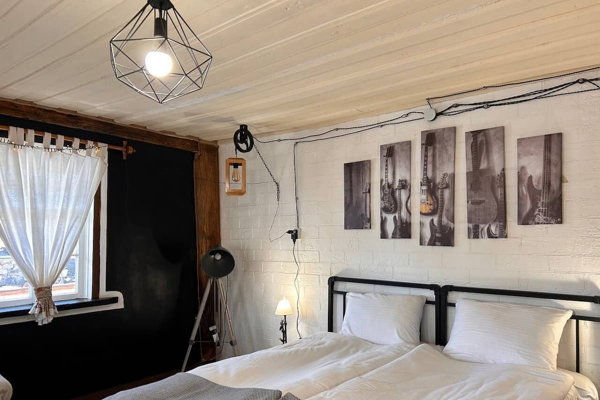 A bedroom with a large bed, a window with sheer curtains, a desk lamp, and wall art.