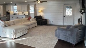 A spacious living room with a large sectional sofa, a gray armchair, and a coffee table. There is a kitchen in the background with white cabinets and a window. - Sleeps 6-8; Across from Sand Mountain Park (Albertville)