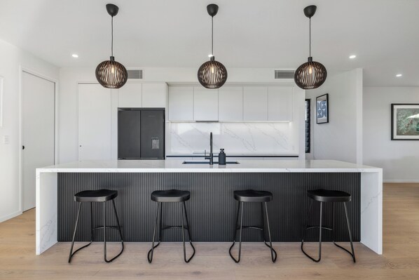 A modern kitchen with a white marble countertop, three black bar stools, and three pendant lights.