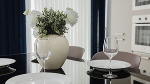 A dining table with white plates and wine glasses, a vase with white flowers, and a modern kitchen in the background. - This apartment is a 1 bedroom(s), 1 bathrooms, located in San Pawl il-Baħar, null. (San Pawl il-Baħar)