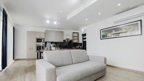 A modern living room with a grey sofa, a kitchen in the background, and a framed picture on the wall. - This apartment is a 1 bedroom(s), 1 bathrooms, located in San Pawl il-Baħar, null. (San Pawl il-Baħar)