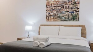 A neatly made bed with white linens and a gray throw blanket, a bedside lamp, and a wall-mounted picture of a riverside scene.